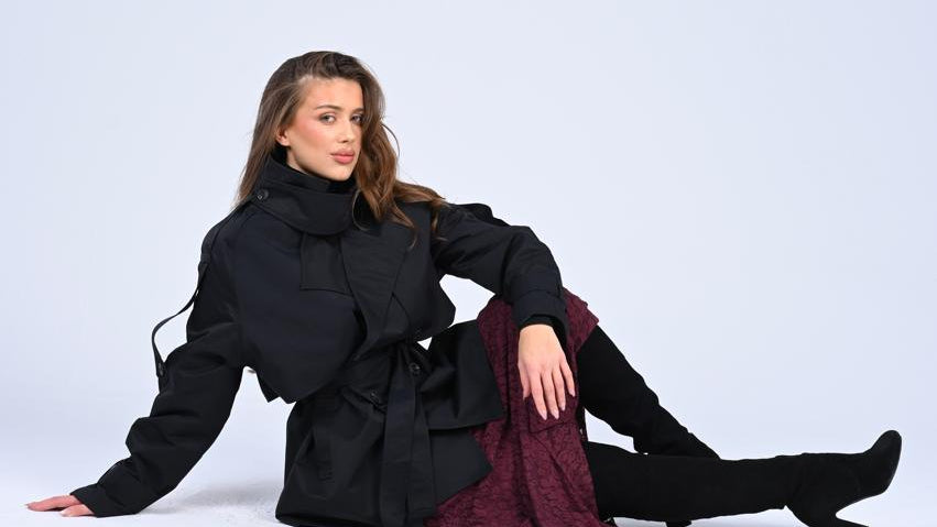 Woman sitting on a white background wearing a black coat and maroon pants.